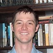 A smiling man with short hair is standing in front of a bookshelf filled with books.