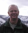A smiling man in a jacket stands outdoors with snowy mountains in the background.