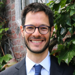 A professional portrait of a smiling man in a suit and tie, standing against a backdrop of greenery and brick.