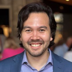 A smiling man with dark hair is wearing a blue suit and a light blue shirt, set against a blurred social gathering background.