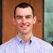 Professional headshot of a smiling man in a light blue shirt, standing in front of a wooden wall.