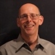 A smiling man with glasses wearing a light grey shirt against a dark background.