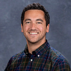 Professional headshot of a smiling man with dark curly hair, wearing a plaid shirt.
