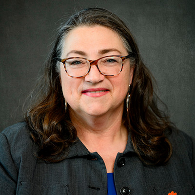 Professional portrait of a woman with long hair wearing glasses and a dark blazer, smiling at the camera.