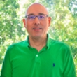 A smiling man wearing a green polo shirt, standing in front of a blurred natural background.