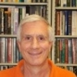 A smiling man wearing an orange shirt stands in front of a bookshelf filled with books.