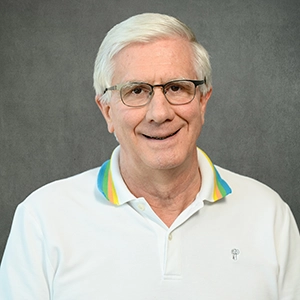 Smiling older man with white hair and glasses wearing a white polo shirt with a colorful striped collar against a gray studio background.