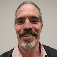 A close-up portrait of a man smiling, with a greying mustache and beard.