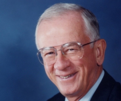 Smiling portrait of a middle-aged man with glasses and a blue suit against a dark background.