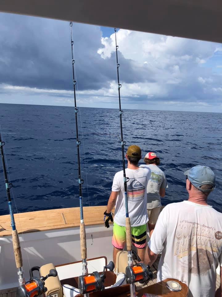 Three fishermen on a boat are prepared to fish, with rods set up and a calm ocean stretching before them under a cloudy sky.