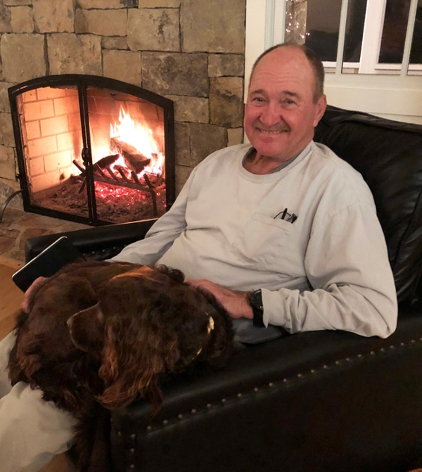 A smiling man sits in a cozy chair by a fireplace, holding a dog in his lap.