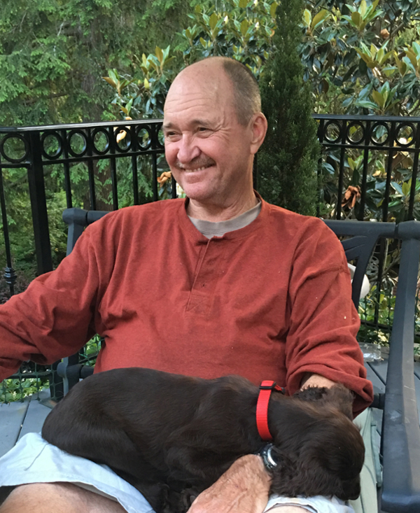 A smiling man in an orange long-sleeve shirt sitting outside with a brown dog resting on his lap.