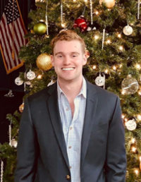 A smiling young man in a dark blazer stands in front of a decorated Christmas tree.