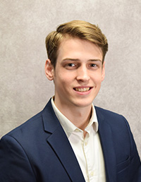 Headshot of a young man in a dark suit and white shirt, smiling against a gray backdrop.