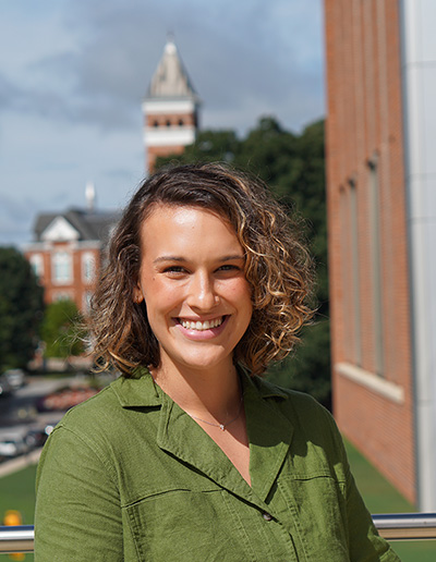 Smiling woman with curly hair wearing a green shirt, with a campus building in the background.