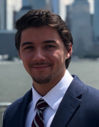 A young man in a suit smiles confidently against an urban backdrop.