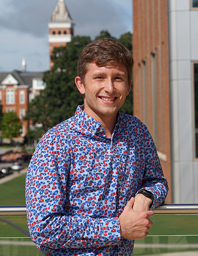 A young man in a floral shirt smiles confidently on a balcony with a campus in the background.