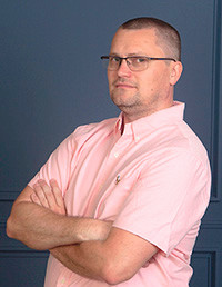 Professional portrait of a man wearing a pink shirt with glasses, standing confidently with arms crossed against a blue background.