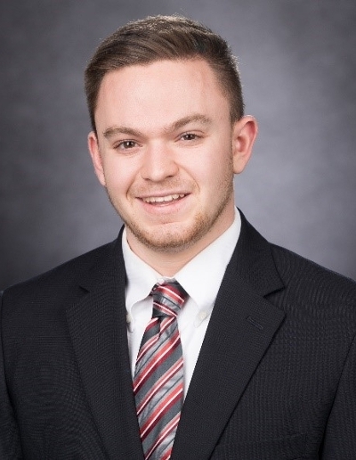 A professional headshot of a young man wearing a black suit, white shirt, and a red and gray striped tie, smiling against a dark gray background.