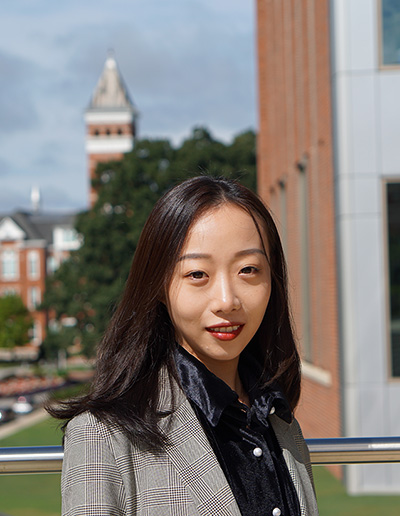 A professional portrait of a woman in a stylish blazer, posing outdoors with a blurred university campus background.