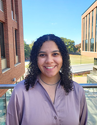 A smiling young woman with curly hair stands outside in front of modern buildings and greenery.