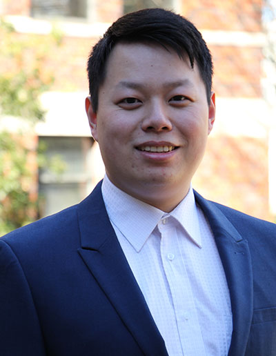 A smiling professional man in a suit, standing outdoors against a blurred background of brick buildings.