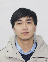 Portrait of a young man with dark hair wearing a light beige jacket over a blue shirt.
