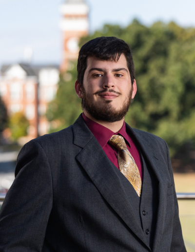 A professional man in a suit and tie poses confidently outdoors, with a campus building and trees in the background.