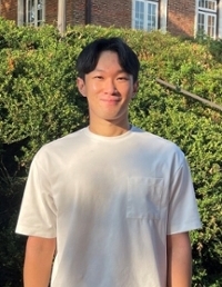 A young man stands outdoors in a white t-shirt, smiling warmly against a backdrop of greenery.
