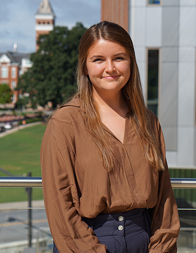 A young woman with long brown hair stands outdoors, wearing a brown blouse and navy pants, with a university campus in the background.