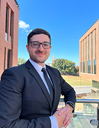 A professional man in a suit poses outdoors with a modern architectural background and a clear blue sky.