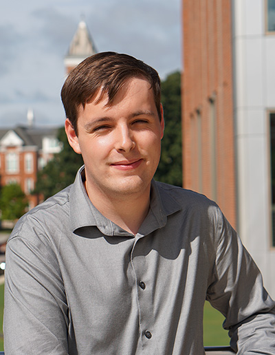 A young man in a gray button-up shirt smiles confidently outdoors with a university campus in the background.