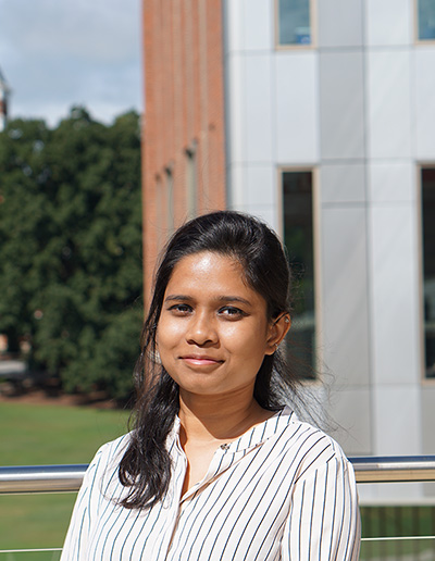 A young woman with long dark hair, wearing a striped shirt, smiles at the camera with a modern building in the background.