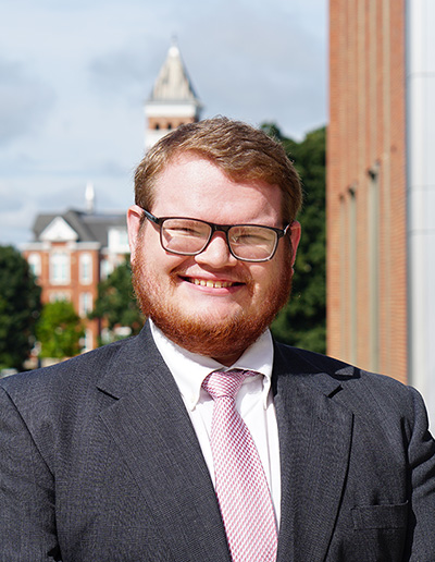 Portrait of a young man with a beard, wearing glasses and a suit, smiling against a backdrop of a university campus.