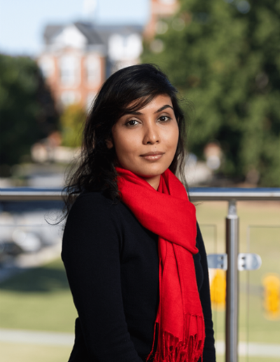 A woman with long dark hair wearing a black sweater and a bright red scarf poses outdoors, with blurred greenery and buildings in the background.