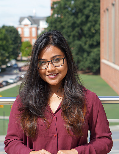 A woman with long black hair and glasses smiles while wearing a burgundy shirt, standing outdoors against a blurred background of green grass and buildings.