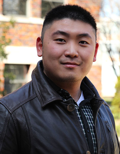 A portrait of a young man wearing a leather jacket and standing outside with a slight smile.