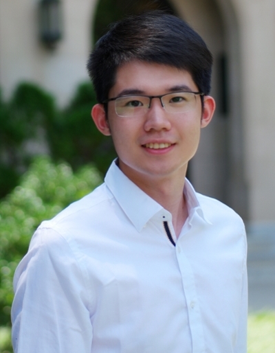 A young man wearing a white shirt and glasses, smiling outdoors in a slightly blurred garden setting.