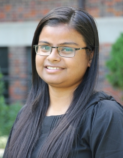 Portrait of a smiling young woman with long black hair and glasses, dressed in a black top, standing outdoors.