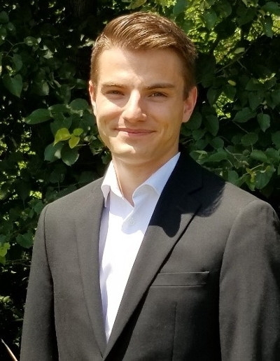 A young man in a black suit and white shirt smiles against a backdrop of green foliage.