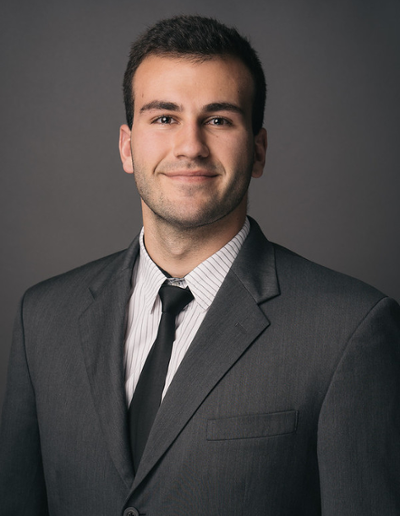 A professional headshot of a young man wearing a black suit, white dress shirt, and black tie, smiling at the camera.