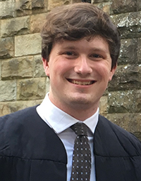A smiling graduate in a cap and gown, posing in front of a stone wall.