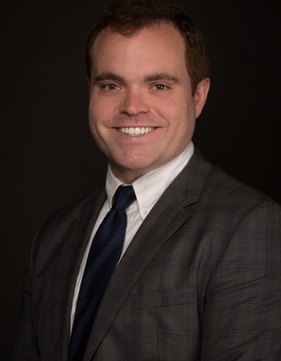 Professional headshot of a smiling man in a suit and tie against a dark background.