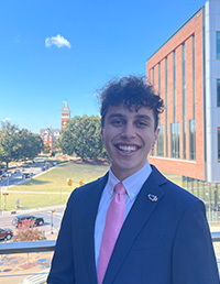 A smiling young man in a suit and tie stands outdoors with a university building and clock tower visible in the background.