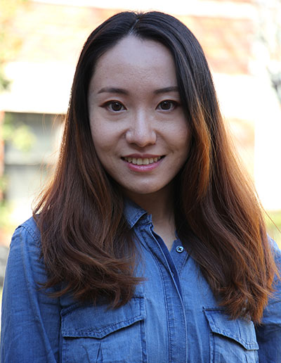 A smiling woman with long, wavy hair wearing a denim shirt, standing outdoors.