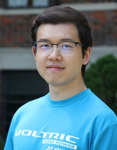 A young man in a blue VOLTRIC sweatshirt wearing glasses, standing outdoors with a blurred building and greenery in the background.