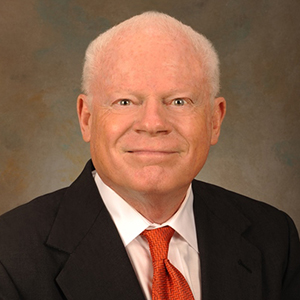 Professional headshot of a man in a black suit with an orange tie and a light-colored shirt, smiling against a neutral background.