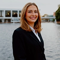 Professional portrait of a smiling woman in a black suit, standing by a water body in an outdoor setting.