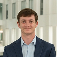Professional headshot of a young man in a suit, smiling in an office environment.