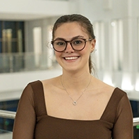 A smiling young woman with glasses wearing a brown top, posing in a modern indoor setting.